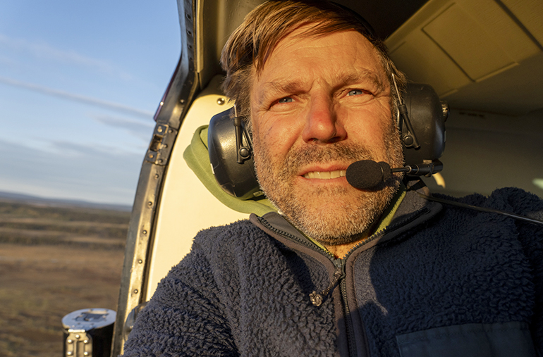 Johan Hammar sitter i en helikopter med öppning ut mot luften. Han har hörlurar på sig och solen lyser i hans ansikte. 