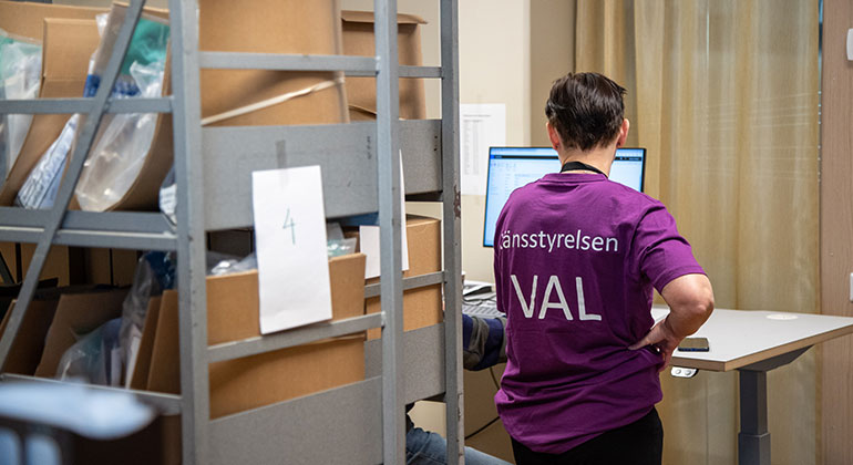 En person står vid en datorskärm. Personen har en lila tröja med texten Länsstyrelsen VAL. I förgrunden en hylla med lådor. 