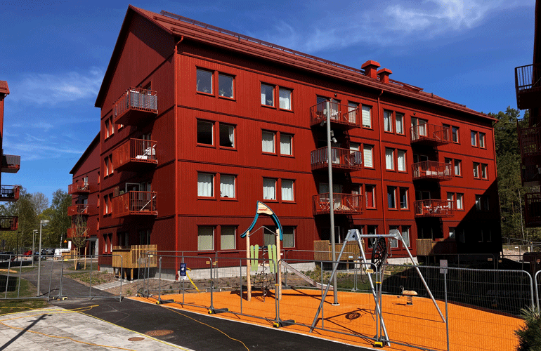 Ett rött flerfamiljshus med balkonger. Framför huset ligger en lekplats med byggstängsel runtom.