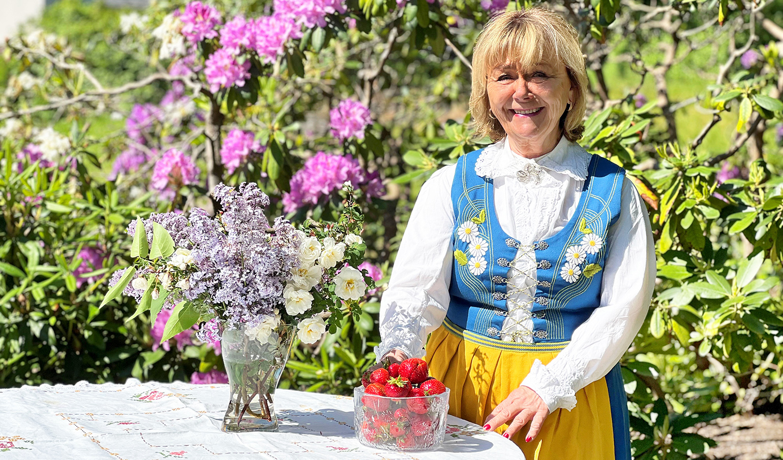 Beatrice Ask står utomhus i solsken, hon har en blågul dräkt på sig. På bordet framför henne står en skål med jordgubbar och en vas med blommor. 