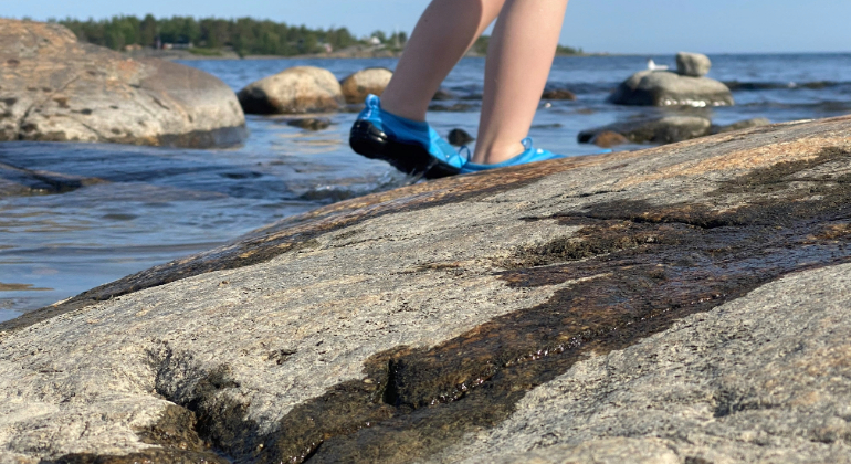 En bild av fötterna av ett barn som går i sandalerna på en sten vid en strand.