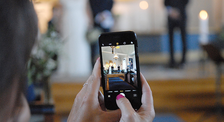 En person håller upp en mobiltelefon och i displayen ser man ett brudpar i en kyrka