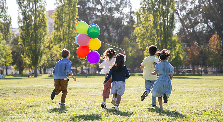 Fem barn som springer på gräset i en park, de håller sex ballonger i färgerna grön, gul, orange, röd, rosa och lila.