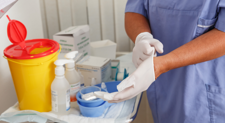 En vårdpersonal sätter på sig plasthandskar. På ett bord står material som exempelvis en behållare för använda sprutor.